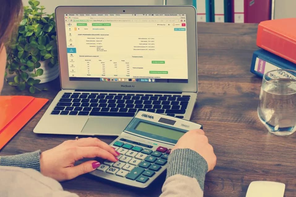 Hands using a calculator beside a laptop showing an account ledger